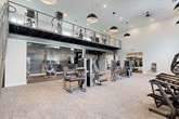 Located at 140 SW 91st Ave in Plantation, Florida, this generously sized fitness facility offers a thoughtfully designed, multi-zone workout environment spanning a single floor with a striking mezzanine level overhead. The main floor is characterized by soaring double-height ceilings, crisp white walls, and patterned carpeted flooring with subtle geometric gold accents, creating a clean, contemporary aesthetic throughout the primary exercise areas.

The main floor is organized into distinct training zones. A large open area accommodates a full array of selectorized strength machines, treadmills, and cardio equipment, bathed in natural light from oversized windows that frame views of the surrounding Florida landscape. Bold dome-shaped pendant lights hang from the elevated ceiling, reinforcing the industrial-modern design language. A dedicated free weights room features rubber flooring, dumbbell racks, a Smith machine, benches, and large wall-mounted mirrors, while a separate functional training area includes a TKO multi-station rig, a heavy bag, and a mini trampoline.

A metal staircase with sleek railings leads to the mezzanine level, where additional cardio and strength equipment is positioned beneath an exposed black structural ceiling. Floor-to-ceiling glass panels along the mezzanine edge overlook the main floor below, enhancing the sense of openness and visual connectivity throughout this well-appointed facility.