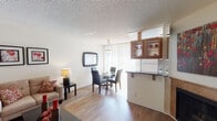 This single-floor space showcases a thoughtfully designed open layout that seamlessly connects the main living areas. The living room is anchored by a tiled fireplace surround and flows naturally into a dedicated dining area, both finished with warm light hardwood flooring throughout. A textured ceiling adds visual depth, while neutral walls provide a versatile backdrop. The living and dining zone benefits from natural light through a large window, enhancing the open, airy atmosphere.

The kitchen is efficiently arranged with rich granite countertops, warm honey-toned cabinetry featuring glass-paneled upper doors, a double stainless steel sink, and tile flooring. Ample cabinet storage and clean lines give the space a practical yet polished character.

A distinctive architectural feature is the semi-private bedroom area, elevated slightly and separated from the main living space by a white decorative railing. This loft-style arrangement creates a sense of defined privacy while maintaining visual connectivity with the rest of the home. The bedroom area is finished with carpeting and includes a ceiling fan. The entry door is positioned near this transition zone, providing a clear and accessible flow throughout the entire single-level layout.