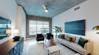 This single-floor residence showcases a compelling mix of industrial and contemporary design elements throughout its well-appointed layout. The living room immediately draws attention with its striking teal-toned textured ceiling, complemented by a ceiling fan with a brass fixture and large windows that frame urban views and fill the space with natural light. Light hardwood flooring flows throughout the common areas, grounding the eclectic aesthetic. A decorative accent wall in raw, oxidized tones adds warmth and texture to the living space, which comfortably accommodates a seating area with a wall-mounted television and a geometric media console.

The kitchen features a large island with granite countertops and bar seating for multiple guests, dark espresso cabinetry, stainless steel appliances including a full-size refrigerator, range, and microwave, and pendant lighting overhead. Both bedrooms carry the industrial aesthetic with textured accent walls, windows with outdoor views, and cozy carpeting. Each bathroom is finished with white subway tile walk-in showers with glass enclosures, granite vanity countertops, white cabinetry, and wood-look flooring, creating a clean, modern feel throughout.