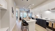 This efficiently designed single-floor residence offers a thoughtful layout across one bedroom, one bathroom, a kitchen, living room, laundry area, and two closets. The kitchen is a standout feature, showcasing bold two-tone cabinetry with navy lower cabinets and crisp white uppers, complemented by a herringbone tile backsplash, stainless steel appliances, a matte black faucet, and a central island with bar seating. Track lighting overhead enhances both function and style. The kitchen flows seamlessly into the living area, where large windows and a glass-panel door flood the space with natural light and provide direct outdoor access. Light hardwood-style flooring runs throughout the main living areas, tying the open-concept space together. The bedroom is a calm retreat featuring a large window with outdoor views, soft carpeting, and ample room for a full furniture arrangement. The full bathroom includes a white shaker-style vanity with quartz countertops, black hardware accents, and direct access to a spacious walk-in closet with built-in shelving and hanging rods. A dedicated laundry area and additional closet storage add practical value. The overall aesthetic is modern and cohesive, with neutral tones, quality finishes, and a clean, contemporary design throughout.