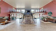 Located at 8301 16 1/2 Mile Rd in Sterling Heights, Michigan, the AVA Sterling Apartment leasing office is a generously scaled, two-story commercial space that makes a bold and memorable first impression. The expansive main floor serves as the centerpiece of the space, featuring soaring ceilings, large floor-to-ceiling windows, and wide-plank hardwood-style flooring that extends throughout. Natural light floods the interior, offering clear views of the surrounding outdoor landscape.

A dramatic dual staircase anchors the heart of the main floor, with sleek metal railings and open treads that create an architectural focal point. The living area is appointed with a tufted leather sofa set against a striking multi-toned reclaimed wood accent wall, beneath the prominently displayed AVA Sterling Apartment signage. An ornate spherical chandelier hangs overhead, complementing the eclectic design aesthetic. A classic red vintage car on display adds a distinctive, eye-catching element to the space.

The entrance features a tall, multi-panel glass curtain wall framing a mixed-material accent wall with an oversized clock, creating a grand and welcoming reception area. The upper level offers additional seating areas with views overlooking the surrounding community, bathed in natural light through generous windows. Warm rose-toned walls and curated decor details throughout lend the space a vibrant, inviting character.