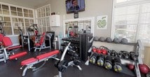 This fitness center is a comprehensively outfitted facility designed to support a full range of workout routines. The space is housed in a bright, single-floor layout with neutral gray walls, white trim, and a dark rubber flooring that is both durable and practical for exercise use. Large windows line multiple walls, flooding the interior with natural light and creating an open, energizing atmosphere. Transom-style windows and glass-paneled interior doors add an architectural charm that distinguishes this facility from a standard gym setting.

The cardio section features multiple treadmills and an elliptical trainer positioned to take advantage of the natural light, alongside a stability ball for supplementary training. A dedicated strength training zone includes several selectorized cable and resistance machines with red upholstered padding, providing a variety of targeted muscle group workouts. A well-stocked dumbbell rack lines one wall, accompanied by adjustable weight benches and a wall-mounted flat-screen television for entertainment during workouts. Additional cable systems and a multi-functional training station round out the strength equipment offerings. A ceiling fan aids in air circulation, and recessed lighting ensures the space remains well-lit throughout. The facility also connects to an adjacent corridor area, suggesting access to additional amenities nearby.