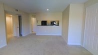 This single-floor space offers a clean, open-concept layout designed for comfortable everyday living. The main living area is finished with wall-to-wall neutral carpeting, creating a warm and cohesive feel underfoot. Smooth, light-toned walls and recessed lighting contribute to a bright, understated ambiance throughout the space.

A defining architectural feature is the pass-through kitchen window, which opens directly from the kitchen into the living area. This design element enhances connectivity between the two spaces while maintaining a clear visual boundary. The kitchen is equipped with wooden cabinetry, dark countertops, a black refrigerator, and a built-in oven, offering functional cooking amenities in a compact, efficient layout.

A hallway visible toward the rear of the living area leads to additional rooms, suggesting a practical floor plan with straightforward circulation. The textured ceiling and recessed lighting add subtle dimension without overwhelming the overall simplicity of the design. The neutral color palette of beige and white throughout lends the space a versatile, move-in-ready quality suitable for a variety of furnishing styles.