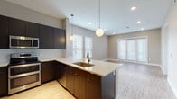 This single-floor space showcases a contemporary design with a cohesive, open layout connecting the kitchen, dining, and living areas. The kitchen is a standout feature, equipped with dark espresso cabinetry, stainless steel appliances including a double oven range, built-in microwave, French door refrigerator, and dishwasher. A white quartz peninsula with an undermount sink doubles as a breakfast bar, illuminated by two globe pendant lights that add a modern aesthetic touch. White subway tile backsplash provides a clean contrast to the dark cabinetry.

The living area benefits from abundant natural light through multiple windows fitted with white blinds, as well as French doors that open to the exterior. Light wood-style flooring runs consistently throughout, enhancing the airy feel of the space. The bedroom is a well-proportioned room with dual windows, a ceiling fan, and ample natural light.

The bathroom features a long white vanity with a mirror, wood-style flooring, and a tub-shower combination adorned with bold marble-style tile surround. Neutral warm-gray walls throughout the unit create a calming, move-in-ready atmosphere well-suited for modern living.