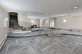 This building lobby in Chilliwack, BC offers a generously proportioned entry space that sets a functional and welcoming tone for residents and visitors alike. The floor is covered in patterned grey carpet tiles laid in a herringbone arrangement, adding a subtle visual texture throughout the open area. A standout architectural feature is the curved staircase with ornate black wrought-iron railings, which adds a decorative touch and serves as a central visual element within the space. The lobby provides access to an elevator, ensuring convenient multi-floor connectivity. Near the main entrance, a textured stone accent wall frames the glass entry doors, which allow natural light to filter in and offer views of the surrounding streetscape. A secondary set of glass doors provides an additional point of entry. Wall-mounted sconce lighting complements the flush ceiling fixtures, maintaining a bright and evenly lit environment. A built-in mailbox unit and a small console table are positioned along the interior walls, supporting everyday resident needs. Baseboard heating runs along the perimeter, ensuring comfort throughout the seasons. The light-toned walls and textured ceiling contribute to the overall clean, neutral aesthetic of this practical and well-appointed lobby.