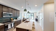 This well-appointed single-floor residence offers a thoughtfully designed open-concept layout that seamlessly connects the kitchen, dining, and living areas. The kitchen is a standout feature, showcasing rich dark-stained cabinetry, a striking teal subway tile backsplash, stainless steel appliances including a side-by-side refrigerator with ice and water dispenser, a gas range, and a microwave. A generously sized central island with a white quartz countertop, an undermount sink, and bar seating serves as both a functional workspace and a gathering spot. Pendant lighting in a muted green tone adds a contemporary accent overhead. Light wood-style flooring flows throughout the main living areas, enhancing the bright, airy atmosphere. The living room is anchored by large black-framed windows and a sliding glass door that fills the space with natural light and provides outdoor access. The primary bedroom features plush carpeting, a large window with outdoor views, and an en-suite bathroom with a walk-in tiled shower and an adjoining walk-in closet. A second bedroom, equally comfortable and well-lit, is served by a full bathroom featuring a tiled tub-shower combination. The overall aesthetic is clean and modern, with a neutral palette punctuated by bold accent details throughout.