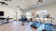 This fitness center offers a thoughtfully designed, single-floor layout that accommodates a comprehensive range of workout options. The space is finished with clean white walls and durable wood-look flooring throughout, creating a bright, contemporary atmosphere enhanced by sleek linear LED lighting and industrial-style ceiling fans.

The cardio zone is generously appointed with multiple treadmills, elliptical trainers, a StairMaster, and a rowing machine, all positioned to take advantage of large windows that frame views of the surrounding outdoor area. A wall-mounted flat-screen television adds entertainment for cardio sessions.

The strength training area is equally well-stocked, featuring a cable machine, multi-function selectorized weight machines, a Smith machine with free weight plates, adjustable benches, and a dumbbell rack. Floor-to-ceiling mirrors amplify the sense of space and allow users to monitor their form throughout their workouts.

A dedicated functional fitness and stretching room includes a warmer hardwood floor, a wall-mounted storage rack holding medicine balls, kettlebells, stability balls, and resistance accessories, alongside another flat-screen display. Decorative tile accent walls in muted blue and earth tones add visual character to this area.

A water fountain and storage cabinetry are also conveniently integrated into the space. Direct access to an outdoor area is provided through glass doors, connecting the fitness center seamlessly to the broader community environment.