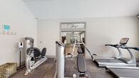 Located at 100 Legacy Way in Yorktown, Virginia, this fitness center is a thoughtfully designed, single-level facility offering a comprehensive workout experience across multiple interconnected zones. The space is divided into distinct areas, each tailored to a specific type of exercise. The strength training area features a robust selection of Hoist cable machines, a lat pulldown station, and a full rack of dumbbells arranged along floor-to-ceiling mirrors that amplify the sense of space. Patterned carpet tiles throughout this zone provide comfort underfoot during weight training sessions. Accessible via elegant French doors with glass transom windows above, the cardio area is outfitted with True-brand treadmills, elliptical trainers, and a recumbent bike. Large windows with slatted blinds flood this room with natural light and offer views of the surrounding greenery, while wall-mounted televisions keep users entertained during workouts. Vaulted ceilings add a sense of openness throughout. A dedicated cycling and group fitness studio features smooth hardwood-style flooring, a row of stationary bikes, full-wall mirrors, ceiling fans for air circulation, a large flat-screen TV, and resistance training accessories mounted to the wall, creating an inviting environment for both individual and group exercise sessions.