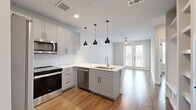 This thoughtfully designed 1-bedroom, 1-bathroom residence offers a modern and functional layout across a single floor. The open-concept kitchen and living area serves as the heart of the home, anchored by a large center island with a white quartz countertop, an undermount sink, and striking matte black pendant lights overhead. The kitchen features soft gray shaker-style cabinetry, a herringbone tile backsplash, stainless steel appliances including a range, over-the-range microwave, and refrigerator, all set against crisp white countertops. Built-in open shelving near the entryway provides practical storage and adds a custom touch. Warm hardwood-style flooring flows throughout the entire space, adding cohesion and comfort. The bedroom is generously sized, bathed in natural light through a large window with views of greenery, and includes a ceiling fan. The full bathroom is well-appointed with a long vanity offering ample storage, a framed mirror, and a glass-enclosed walk-in shower. A dedicated closet with built-in shelving provides convenient organization directly off the bathroom. The overall aesthetic is clean, contemporary, and move-in ready.