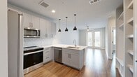 This thoughtfully designed 1-bedroom, 1-bathroom residence offers a modern and functional layout across a single floor. The open-concept kitchen and living area serves as the heart of the home, anchored by a large center island with a white quartz countertop, an undermount sink, and striking matte black pendant lights overhead. The kitchen features soft gray shaker-style cabinetry, a herringbone tile backsplash, stainless steel appliances including a range, over-the-range microwave, and refrigerator, all set against crisp white countertops. Built-in open shelving near the entryway provides practical storage and adds a custom touch. Warm hardwood-style flooring flows throughout the entire space, adding cohesion and comfort. The bedroom is generously sized, bathed in natural light through a large window with views of greenery, and includes a ceiling fan. The full bathroom is well-appointed with a long vanity offering ample storage, a framed mirror, and a glass-enclosed walk-in shower. A dedicated closet with built-in shelving provides convenient organization directly off the bathroom. The overall aesthetic is clean, contemporary, and move-in ready.