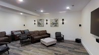 This single-floor space presents a thoughtfully designed lounge area that balances comfort and contemporary style. The room is anchored by a rich, dark brown leather sofa accompanied by matching accent chairs, all arranged around a tufted white ottoman that serves as a central coffee table. The seating arrangement encourages conversation and relaxation in equal measure.

The walls are finished in a clean, neutral white that enhances the sense of openness and brightness throughout the space. Recessed ceiling lights provide even, ambient illumination, while a series of sleek vertical wall sconces add a refined decorative touch. Two framed abstract artworks adorn the walls, introducing subtle pops of color and visual interest against the otherwise minimalist backdrop.

Underfoot, a multi-toned patterned carpet in earth and cool tones adds texture and warmth to the space, complementing the dark furniture and white walls cohesively. The ceiling features a subtle architectural stepped detail, adding dimension without overwhelming the room's clean aesthetic.

Overall, the space exudes a polished, modern ambiance well-suited for a communal lounge, waiting area, or gathering space, offering a welcoming environment that is both functional and visually appealing.