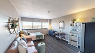 Located at 14703 Park Row Boulevard in Houston, TX, this efficiently designed one-bedroom, one-bathroom unit makes excellent use of its compact footprint across a single floor. The entrance corridor leads directly into the main living area, where striped carpet flooring and a textured ceiling set the tone throughout. Large windows line the exterior wall, flooding the space with natural light and offering open views of the surrounding neighborhood skyline.

The main room functions as a combined living and sleeping area, with ample room for a full-sized bed, seating, and a dining arrangement. A kitchenette along one wall features white cabinetry, a subway tile backsplash, stainless steel refrigerator, microwave, and a single-basin sink — providing essential cooking and storage functionality within a streamlined design.

The bathroom is accessed via a separate door and features a long floating vanity with an illuminated mirror, light tile flooring, and a shower-tub combination with curtain. A dedicated closet provides convenient storage just off the main living space.

The unit's layout is thoughtfully organized to maximize every square foot, with natural light and exterior views enhancing the overall sense of openness throughout.