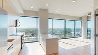 Located in the heart of Raleigh, NC, this high-rise residence at 9 Harrington St offers a sleek, modern design across a thoughtfully organized single floor. The open-concept living area is defined by dramatic floor-to-ceiling windows that flood the space with abundant natural light and frame sweeping panoramic views of the Raleigh skyline and the lush tree canopy stretching across the horizon. Light-toned hardwood flooring runs throughout, complementing the clean, contemporary aesthetic.

The kitchen is well-appointed with light wood cabinetry, a classic white subway tile backsplash, white countertops, and a full suite of stainless steel appliances, including a French door refrigerator, dishwasher, range, and microwave. A center island provides additional prep and serving space.

Six bedrooms offer versatile accommodations, with several rooms featuring direct access to a private balcony and their own floor-to-ceiling glass walls showcasing city and treetop views. The three bathrooms are finished in a modern style with light wood vanities, white countertops, and clean chrome fixtures.

Additional highlights include a dedicated laundry area, wire-shelf closets with generous storage, and two hallways that provide efficient flow throughout. The entrance leads seamlessly into the main living areas, making this an exceptionally functional and visually striking residence.