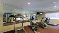 This single-floor fitness center offers a dedicated space for cardiovascular exercise and general wellness. The room is outfitted with a selection of cardio equipment, including treadmills, elliptical trainers, and a stationary bike, providing users with a variety of workout options. A large wall mirror spans a significant portion of one wall, enhancing the sense of openness and allowing users to monitor their form during exercise. Recessed lighting is evenly distributed across the textured ceiling, ensuring the space is well-lit throughout. A wall-mounted flat-screen television is positioned for easy viewing from the equipment, adding an element of entertainment to workouts. Natural light filters in through windows along the walls, contributing to a bright and welcoming atmosphere. The flooring features dark carpeting, which adds a layer of comfort underfoot. Neutral-toned walls complement the overall clean and functional aesthetic of the room. An emergency exit sign and fire safety equipment are also present, reflecting attention to safety standards. Overall, this fitness center is a practical and well-maintained space designed to support an active lifestyle.