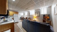 This fully finished lower-level suite offers a compact yet well-organized layout across a single floor, comprising a combined living and dining area, a kitchen, a bedroom, and a full bathroom. The open main living area features durable hardwood-style flooring that transitions to plush carpet, and is topped throughout by a distinctive patterned drop-tile ceiling. Multiple above-grade windows fitted with vertical blinds allow natural light to filter in, brightening the interior. The living area is configured to accommodate both a seating arrangement and a dining table with chairs, making efficient use of the available space. A television viewing area with a recliner and sofa creates a comfortable lounge zone. The kitchen is outfitted with warm oak cabinetry, light countertops, a stainless steel sink, a full-size refrigerator, a dishwasher, and a coffee station — providing all the essentials for everyday living. The bedroom is carpeted and features a window with vertical blinds, a dresser, and flanking nightstands, offering a quiet and functional retreat. Adjacent to the bedroom, the bathroom includes a wood-vanity cabinet, a tub-shower combination with a decorative curtain, and hardwood-style flooring, completing this self-contained lower-level suite.