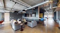 This single-floor communal space showcases a refined industrial aesthetic, blending raw architectural elements with thoughtfully curated furnishings. Exposed concrete ceilings with visible ductwork and structural columns set the tone throughout, while polished concrete floors unify the open, expansive layout.

The lounge area serves as a central focal point, anchored by a dramatic darkened-metal feature wall housing a linear fireplace and mounted flat-screen televisions. Comfortable grey seating arrangements are complemented by bold pops of color, including vibrant orange armchairs and a striking blue Eames-style bar stool. Pendant lighting adds warmth to the industrial palette.

A distinct booth-style seating section features warm wood-paneled walls and intimate café tables, creating a semi-private nook for smaller gatherings. Adjacent to this is a well-appointed kitchen bar with white cabinetry, a patterned tile backsplash, and generous counter seating.

A games area includes a pool table set beneath a bold pendant light, surrounded by mixed flooring materials including wood planks and polished concrete. Throughout the space, barn-style sliding doors, exposed concrete columns, and curated artwork contribute to the cohesive, modern-industrial ambiance.