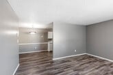 This single-floor unit offers a practical and well-organized layout designed for comfortable everyday living. The interior is finished with warm, neutral gray tones on the walls and complemented by durable wood-look flooring that runs throughout the main living areas and kitchen, creating a cohesive and easy-to-maintain aesthetic. Textured ceilings are present throughout the space.

The living area is open and spacious, featuring a large window with vertical blinds that allows natural light to filter in and provides a view of the outdoors. It flows seamlessly toward the kitchen and dining area, making the space feel connected and functional. The kitchen is well-equipped with ample white cabinetry, light-toned countertops, a built-in sink, a dishwasher, a black refrigerator, and a range, offering generous storage and prep space. A pendant light fixture anchors the dining area adjacent to the kitchen.

The two bedrooms are carpeted in a neutral tan tone, each featuring a window with horizontal blinds that brings in natural light. Both rooms offer a quiet, comfortable retreat with sufficient space for furnishings.

The bathroom includes a single-sink vanity with white cabinetry, a large mirror with overhead lighting, and a shower/tub combination, all finished with the same wood-look flooring found throughout the unit.