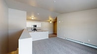 This single-floor space is designed with an open-concept layout that seamlessly connects the kitchen and living area, creating a functional and cohesive environment. The kitchen is equipped with stainless steel appliances, including a microwave and range, and features white cabinetry that provides ample storage. The countertops display a granite-style finish, complementing the clean, neutral color palette found throughout the space. A peninsula counter extends into the living area, offering additional workspace or casual seating and serving as a natural divider between the two zones.

The living area is finished with plush wall-to-wall carpeting in a neutral gray tone, contributing to a comfortable and quiet atmosphere. Vaulted ceilings in the living zone add a sense of openness and vertical interest, while a ceiling fan provides both airflow and ambient lighting. A baseboard heater runs along the exterior wall, indicating an efficient heating solution. The walls throughout are painted in a warm beige tone, lending a cohesive and inviting feel. Recessed and flush-mount lighting fixtures illuminate both areas, ensuring a well-lit interior.