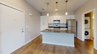 Located at 14847 Piper Lakes Cir in Grand Haven, Michigan, this one-bedroom, one-bathroom unit offers a thoughtfully designed single-floor layout that maximizes its compact footprint. The open-concept living and kitchen area forms the heart of the space, featuring warm wood-look flooring throughout and neutral taupe-toned walls that create a clean, modern aesthetic. The kitchen is well-appointed with white shaker-style cabinetry reaching toward the ceiling for ample storage, dark countertops, a center island with a built-in sink, and a full suite of stainless steel appliances including a range, over-the-range microwave, and refrigerator. Three pendant lights with gold-tone fixtures hang above the island, adding a contemporary touch. The living area receives natural light through a large window fitted with white blinds. The bedroom is comfortably sized, finished with plush carpeting, a window overlooking the outdoors, and a built-in closet with sliding doors for convenient storage. A hallway leading to the bathroom also houses a stacked washer and dryer unit, providing in-unit laundry convenience. The full bathroom includes a shower-tub combination. Throughout, the space maintains a cohesive, neutral palette with clean white trim and consistent flooring, offering a move-in-ready feel.