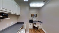 The business center is a thoughtfully designed workspace that maximizes functionality within a compact footprint. The room features built-in workstations with smooth gray countertops extending along two walls, providing generous surface area for productivity. White cabinetry above and below the desks offers substantial storage, keeping the workspace organized and efficient. Two ergonomic white office chairs are positioned at the workstations, accommodating multiple users simultaneously. A printer is conveniently stationed on the desk, alongside a cork bulletin board mounted on the wall for notes and announcements. The space is illuminated by a large overhead fluorescent panel light, as well as a flush-mount ceiling fixture, ensuring bright, consistent lighting throughout. Crown molding adds a refined architectural detail to the upper perimeter of the room. The warm-toned hardwood flooring contrasts pleasantly with the neutral gray and white color palette of the cabinetry and walls, contributing to a clean, professional atmosphere. Overall, the business center is designed to support a range of office tasks in a tidy, well-appointed environment.
