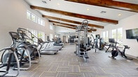 Located in Kissimmee, FL, this generously sized fitness center offers a comprehensive workout environment designed to accommodate a wide variety of exercise routines. The space makes an immediate architectural impression with its soaring vaulted ceilings adorned with bold, rustic wood beams that run the length of the room, creating a warm and distinctive aesthetic against the bright white walls. Large windows and an upper clerestory line the perimeter, flooding the interior with natural light and providing views of the surrounding outdoor greenery and pool area.

The main exercise floor is finished with durable patterned carpet tiles in a neutral gray palette with yellow accents, providing both comfort underfoot and a modern visual appeal. Multiple ceiling fans are suspended throughout, ensuring consistent airflow across the entire space.

The cardio section features a generous row of treadmills and elliptical trainers positioned along the windowed walls, complemented by stationary and spin bikes. The strength training area is equally well-equipped, offering cable machines, multi-station weight systems, free weights, and a dedicated functional training zone with a punching bag, stability balls, kettlebells, and adjustable benches. A wall-mounted television adds convenience for entertainment during workouts. The overall ambiance is bright, open, and well-organized, supporting an energizing fitness experience.