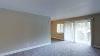 This single-floor residence offers a clean, functional layout with a welcoming and understated aesthetic. The living area is finished with plush wall-to-wall carpeting in a soft gray tone, creating a comfortable and cohesive feel underfoot. Neutral warm beige walls complement the light color palette throughout, lending the space a bright and airy atmosphere.

Natural light is a key feature, with a horizontal window fitted with blinds allowing controlled daylight into the room. A full-height sliding glass door, dressed with sheer vertical blinds, provides additional light and direct access to the outdoors, enhancing the open feel of the interior. The door arrangement suggests the potential for a patio or exterior area just beyond.

A wall-mounted air conditioning unit beneath the window ensures year-round climate control. The textured white ceiling adds subtle visual interest while maintaining a clean overhead finish. Crisp white baseboards frame the perimeter of the room, providing a polished contrast against the neutral walls and carpet.

The open floor plan of the living space offers flexibility for a variety of furniture arrangements, making it easy to configure for different lifestyle needs. Overall, the space presents a straightforward, move-in-ready environment with practical features and a calm, neutral design sensibility.