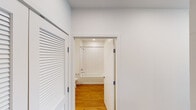 This single-floor unit offers a well-designed layout with a contemporary aesthetic defined by crisp white walls, warm hardwood flooring, and recessed lighting throughout. The open-concept living and kitchen area is a central highlight, where the kitchen is outfitted with flat-front cabinetry in a soft gray finish, stainless steel appliances including a dishwasher, range, and refrigerator, a clean white countertop, and a undermount sink. A sliding barn door adds a modern architectural detail to the space.

The living area is generously proportioned, bathed in natural light through large floor-to-ceiling windows and a glass door that frame expansive urban views of the surrounding cityscape. The bedroom features neutral carpet and bright white walls, providing a quiet, comfortable retreat.

The bathroom is cleanly finished with white subway tile surrounding a full soaking tub and shower combo, a white vanity with an undermount sink, a large mirror, and hardwood flooring that continues from the main living areas. A hallway with a louvered closet door connects the main rooms, completing a functional and cohesive floor plan suited to modern urban living.