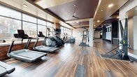 Located at 4905 E Colfax in Denver's 80220 neighborhood, this well-appointed fitness center offers a spacious, single-floor layout designed to support a comprehensive workout experience. The exercise room is thoughtfully zoned to accommodate a wide variety of training modalities. A dedicated cardio area lines the perimeter near large, floor-to-ceiling windows framed by exposed brick columns, flooding the space with natural light and providing open views of the surrounding streetscape. This zone is equipped with treadmills, stair climbers, ellipticals, and stationary bikes, with wall-mounted televisions adding to the amenity. The strength training section features a full dumbbell rack with weight benches, a cable/pulley machine system, and a power rack with a wooden lifting platform. A separate functional training area includes TRX suspension systems and open floor space for stretching or bodyweight exercises, complemented by large mirrors and patterned accent wallpaper. The flooring transitions between rich hardwood-style planks and rubber matting to suit each training zone. Coffered ceilings with recessed lighting and ceiling fans add an architectural refinement throughout, while dark wood paneling and brick accents lend the space a polished, modern-industrial ambiance.