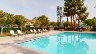 Located in Laguna Niguel, California, this two-level outdoor community amenity space offers a resort-style experience designed for relaxation and recreation. The grounds are anchored by two generously sized swimming pools with crystal-clear blue water, each surrounded by expansive pool decks furnished with lounge chairs, making them ideal for sunbathing and leisure. Complementing the pools are two bubbling spas, one of which is tucked beneath a pergola-style shade structure and flanked by contemporary white lounge chairs, creating a private retreat atmosphere. Lush mature trees, including palms and shade trees, are thoughtfully integrated throughout the landscape, providing natural beauty and a cooling canopy. Outdoor dining areas with umbrella-shaded tables and chairs offer comfortable spaces for socializing, while dual stainless steel BBQ grills provide a premium outdoor cooking station set against a backdrop of manicured greenery and decorative fencing. The space also features a tiled outdoor shower station finished in warm earth tones with Spanish-influenced architectural detailing, including stucco walls and a clay tile roof accent. The overall ambiance is relaxed and upscale, reflecting the Southern California lifestyle with a well-maintained, community-focused design.