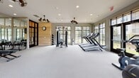Located at 1100 Eagle Hall Ln in Goose Creek, South Carolina, this fitness center is a well-appointed, single-floor facility designed to support a comprehensive workout experience. The space is defined by an open, airy layout with light gray carpeted flooring and a neutral color palette that creates a clean, modern atmosphere throughout.

Large, floor-to-ceiling windows line multiple walls, flooding the interior with natural light and offering views of the surrounding landscaping and community exterior. Black-framed glass doors provide additional connectivity to the outdoors. Decorative dual-blade ceiling fans are positioned throughout, contributing both to the aesthetic character and ventilation of the space.

The fitness center is equipped with a full range of workout stations. A dedicated cardio zone features multiple treadmills, elliptical machines, and rowing equipment, each positioned to take advantage of the outdoor views. A strength training area includes a cable resistance machine, adjustable benches, and a full rack of kettlebells and dumbbells organized along a mirrored wall. The mirrored section also houses mounted flat-screen televisions for entertainment during workouts. A natural wood accent wall with a wall clock serves as a warm focal point, balancing the sleek equipment and reflective surfaces. Recessed lighting supplements the natural light, ensuring the space remains bright and functional at all hours.
