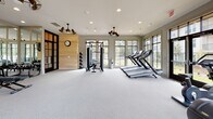 Located at 1100 Eagle Hall Ln in Goose Creek, South Carolina, this fitness center is a well-appointed, single-floor facility designed to support a comprehensive workout experience. The space is defined by an open, airy layout with light gray carpeted flooring and a neutral color palette that creates a clean, modern atmosphere throughout.

Large, floor-to-ceiling windows line multiple walls, flooding the interior with natural light and offering views of the surrounding landscaping and community exterior. Black-framed glass doors provide additional connectivity to the outdoors. Decorative dual-blade ceiling fans are positioned throughout, contributing both to the aesthetic character and ventilation of the space.

The fitness center is equipped with a full range of workout stations. A dedicated cardio zone features multiple treadmills, elliptical machines, and rowing equipment, each positioned to take advantage of the outdoor views. A strength training area includes a cable resistance machine, adjustable benches, and a full rack of kettlebells and dumbbells organized along a mirrored wall. The mirrored section also houses mounted flat-screen televisions for entertainment during workouts. A natural wood accent wall with a wall clock serves as a warm focal point, balancing the sleek equipment and reflective surfaces. Recessed lighting supplements the natural light, ensuring the space remains bright and functional at all hours.
