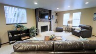 This single-floor space is designed with both comfort and style in mind, centered around a stunning stacked-stone fireplace that serves as the focal point of the living area. The fireplace surround features natural ledger stone in warm, neutral tones, complemented by a bold dark wood accent wall with a chevron-patterned design that extends to the ceiling and frames a wall-mounted flat-screen television. Built-in corner shelving on either side of the fireplace provides display space for decorative accents, adding character to the room. The living area is generously proportioned, accommodating a plush leather sectional sofa and matching leather chairs with an ottoman, arranged around a rustic wood coffee table. Soft, neutral-toned carpeting flows throughout the space, contributing to its warm ambiance. Recessed lighting in the ceiling provides even illumination, while multiple windows dressed with vertical blinds allow natural light to filter into the room. Lush indoor greenery adds a fresh, organic element to the space. The overall aesthetic blends rustic and contemporary design elements, creating an inviting environment that feels both casual and well-appointed.