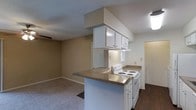 This single-story space features an open-concept design that integrates the kitchen and living area into a cohesive, functional layout. The kitchen is positioned at the heart of the space, anchored by a peninsula counter with a dark speckled laminate surface that doubles as a breakfast bar or prep area. White cabinetry lines the kitchen, including upper cabinets mounted beneath a soffit that extends from the ceiling, providing ample storage. A white electric range with coil burners and a white oven are set against the kitchen wall, complemented by an under-cabinet range hood above. The countertops extend along the kitchen wall, offering generous workspace. Dark hardwood-style flooring runs through the kitchen zone, creating a clear visual distinction from the carpeted living area. The adjoining living space features neutral beige walls and wall-to-wall carpet, contributing to a warm, understated ambiance. A ceiling fan is partially visible in the living area, adding a practical comfort feature. Natural light filters in through a window near the kitchen, while overhead lighting illuminates the space. A hallway visible beyond the kitchen suggests additional rooms beyond the main living area.