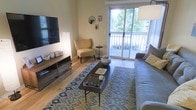 This single-floor residence offers a thoughtfully designed living space that balances comfort and modern style. The living room serves as the heart of the home, featuring warm hardwood flooring that runs throughout and creates a cohesive, inviting atmosphere. A large sliding glass door floods the room with natural light and provides seamless access to a private balcony, where lush greenery is visible beyond the railing, enhancing the sense of a peaceful, nature-connected retreat.

The living area is well-proportioned, comfortably accommodating a plush sofa, a cushioned accent chair, and a sleek, dark wood coffee table at the center. A patterned area rug in muted tones of grey and gold anchors the seating arrangement, adding texture and visual warmth to the space. A low-profile media console along one wall provides practical storage while supporting a wall-mounted flat-screen television. Soft, neutral wall tones complement the warm wood accents and allow the natural light to feel expansive throughout the room.

Decorative wall mirrors and framed accents add personality without overwhelming the clean aesthetic. Overall, the space presents a welcoming, functional layout with a modern sensibility, making excellent use of natural light and outdoor connectivity.