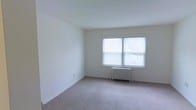 This single-floor space presents a clean, minimalist interior defined by neutral white walls and wall-to-wall carpeting in a warm, light tone. The main room is an open, uncluttered area that feels airy and functional. A prominent double-pane window with horizontal blinds allows an abundance of natural light to filter into the room, creating a bright and welcoming atmosphere. Positioned beneath the window is a classic cast-iron radiator, indicative of a traditional heating system that provides reliable warmth. The smooth white ceiling and matching white trim and baseboards lend the space a cohesive, understated aesthetic. The carpeted flooring adds a sense of comfort underfoot, while the simplicity of the room's design makes it highly adaptable to a variety of furnishing styles and uses. The overall ambiance is calm and neutral, offering a blank canvas that is well-suited for customization. The space is straightforward in its layout, making it easy to navigate, and the ample natural light from the window ensures the interior feels open and inviting throughout the day.