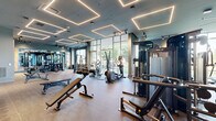 Located at 900 Second Ave N in Nashville, TN, this single-floor fitness facility offers a comprehensive workout experience across two distinct exercise rooms. The space is entered through a sleek vestibule with marble-style tile flooring, leading to glass-paneled double doors branded with the Peloton Studio logo, immediately setting a polished, modern tone.

The Peloton Studio features light hardwood flooring, deep charcoal-gray walls, and a wall of mirrors that amplify the open feel of the room. Track lighting runs the length of the ceiling, and Peloton bikes alongside resistance training equipment are neatly arranged for group or individual use.

The main fitness room is an expansive, well-appointed gym with herringbone-patterned carpet flooring and a bold teal ceiling accented by striking geometric LED light fixtures. Large floor-to-ceiling windows along one wall flood the space with natural light and offer outdoor views. The room is thoroughly equipped with treadmills, cable machines, strength training stations, free weights, benches, and cardio equipment, including air bikes. Ceiling fans provide airflow throughout. Full-length mirrors and warm wood-panel accents add visual depth, while built-in shelving provides organized storage. The overall ambiance is sleek, energizing, and purpose-built for a premium fitness experience.