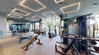 Located at 900 Second Ave N in Nashville, TN, this single-floor fitness facility offers a comprehensive workout experience across two distinct exercise rooms. The space is entered through a sleek vestibule with marble-style tile flooring, leading to glass-paneled double doors branded with the Peloton Studio logo, immediately setting a polished, modern tone.

The Peloton Studio features light hardwood flooring, deep charcoal-gray walls, and a wall of mirrors that amplify the open feel of the room. Track lighting runs the length of the ceiling, and Peloton bikes alongside resistance training equipment are neatly arranged for group or individual use.

The main fitness room is an expansive, well-appointed gym with herringbone-patterned carpet flooring and a bold teal ceiling accented by striking geometric LED light fixtures. Large floor-to-ceiling windows along one wall flood the space with natural light and offer outdoor views. The room is thoroughly equipped with treadmills, cable machines, strength training stations, free weights, benches, and cardio equipment, including air bikes. Ceiling fans provide airflow throughout. Full-length mirrors and warm wood-panel accents add visual depth, while built-in shelving provides organized storage. The overall ambiance is sleek, energizing, and purpose-built for a premium fitness experience.
