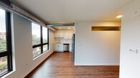 This single-floor unit showcases a thoughtfully designed open-concept layout that seamlessly connects the living area and kitchen. The living space is bathed in natural light through a generous bank of floor-to-ceiling windows framed in dark trim, offering views of the surrounding neighborhood. An exposed spiral ductwork element running along the ceiling adds an industrial character to the otherwise clean, modern aesthetic. Rich hardwood-style flooring flows throughout the main living areas, grounding the space with warmth.

The kitchen is efficiently laid out and well-equipped with white cabinetry, quartz countertops, and a classic white subway tile backsplash. Stainless steel appliances — including a refrigerator, electric range, over-the-range microwave, and dishwasher — complete the functional and stylish cooking space. A small pass-through window connects the kitchen zone to an adjacent area, adding an interesting architectural detail.

The bathroom is clean and contemporary, featuring large-format tile flooring, a dark-cabineted vanity with a backlit LED mirror, and a glass-enclosed walk-in shower. The bedroom is a bright, neutral space with hardwood-style flooring, white walls, and an open closet with a hanging rod, providing practical storage. Throughout, the unit maintains a cohesive, modern palette.