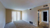 This single-floor residence offers a straightforward and functional layout designed for comfortable everyday living. The main living area is finished with wall-to-wall carpeting in a neutral tone, providing a warm and understated foundation throughout the space. Beige-toned walls contribute to a clean, open atmosphere, while white trim and baseboards frame the room with a classic, tidy finish. A prominent sliding glass door fitted with vertical blinds serves as the primary source of natural light, allowing brightness to fill the living area while offering privacy and light control. HVAC vents integrated into the walls and floors indicate a central air and heating system for year-round climate comfort. A ceiling-mounted ventilation register further supports the home's climate infrastructure. The transition from carpeted living space to what appears to be a tiled or hard-surface floor near the entry suggests distinct functional zones within the home. An interior door visible toward the rear of the space provides access to additional rooms. Overall, the space presents a no-frills, move-in-ready environment with practical features suited for a variety of living arrangements.