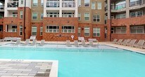 This amenity space is set within a multi-story residential building and offers a compelling combination of outdoor recreation and indoor fitness facilities. The centerpiece is a generously sized outdoor swimming pool with clear, turquoise water, enclosed by the building's striking brick and panel facade rising several stories on multiple sides. The pool deck is lined with rows of lounge chairs, providing ample space for sunbathing and relaxation. Entry points with safety rails offer easy access to the water, and the surrounding hardscape creates a clean, resort-style atmosphere.

Adjacent to the pool area, a fitness center is accessible via a glass-panel entry that allows natural light to flow in while maintaining views of the outdoor courtyard. The gym features an industrial-modern aesthetic, with exposed concrete ceilings, exposed brick accent walls, and bold pendant lighting. The space is comprehensively equipped with a full row of treadmills and cardio machines, cable strength systems, free weights, and a variety of resistance equipment. Large windows connect the interior to the outdoor pool deck, creating a bright and energizing workout environment. Together, these amenities offer a well-rounded and thoughtfully designed lifestyle experience.
