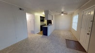 This single-floor unit presents an open-concept layout that seamlessly connects the kitchen and living areas. The kitchen is equipped with dark cabinetry, a wall-mounted range hood, and a built-in peninsula that doubles as a casual dining or prep space. Dark appliances complement the cabinetry, while the peninsula's contrasting navy blue base adds a distinctive design element. A dedicated laundry or utility area is visible adjacent to the kitchen, enclosed behind a white door for a clean, organized appearance.

The living area is finished with wall-to-wall carpet in a neutral tone, providing a comfortable and cohesive feel throughout the space. Crown molding lines the upper edges of the walls, adding a touch of architectural detail. A ceiling fan with a light fixture is centrally positioned in the living area, ensuring year-round air circulation. Natural light filters in through a window along the side wall, brightening the space. The textured popcorn ceiling and white walls create a neutral backdrop, making the unit versatile and easy to personalize.