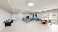 Located at 8600 S Course Dr in Houston, TX, this single-floor space offers a well-designed, open-concept layout ideal for gathering and recreation. The interior is finished with large-format light-toned tile flooring throughout, neutral beige walls, and flush-mounted skylight-style ceiling panels that flood the space with bright, even illumination.

The living area is comfortably appointed with a generous blue sectional sofa and coordinating armchairs arranged around a sleek coffee table, anchored by a textured area rug. A flat-screen TV on a warm wood media console and bold abstract wall art contribute a modern, stylish aesthetic to this lounge zone.

Adjacent to the lounge is a recreational area featuring a full-length shuffleboard table, providing an engaging entertainment option. The space flows seamlessly into a dining area furnished with a round wood table and upholstered chairs, bathed in natural light from large sliding glass doors that offer views of the surrounding outdoor area.

A kitchenette with white cabinetry and a counter workspace supports the dining zone, making this space well-suited for events and social gatherings. Clean lines, an open floor plan, and a cohesive neutral palette give the space a bright, modern, and welcoming atmosphere throughout.