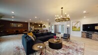 This thoughtfully designed single-floor community space offers a rich variety of gathering areas, each with its own distinct character and purpose. The main lounge is anchored by a generously sized navy sectional sofa paired with round wood and black drum coffee tables, all set beneath a striking brass candelabra chandelier. Recessed lighting and warm wall sconces complement the light hardwood flooring and patterned area rug, creating an inviting atmosphere. Adjacent to the lounge, the kitchen features sleek dark cabinetry, a granite tile backsplash, dark countertops, stainless steel appliances, and a breakfast bar with rich burgundy leather stools beneath elegant arched pendant lights. A separate bar area showcases deep-toned hardwood floors, a decorative geometric-paneled island, high-back bar stools, and industrial cage pendant lighting, with large windows that bring in natural light and views of the outdoors. A cozy lounge nook with red accent chairs flanks a rustic wood-clad structural column fitted with a wall-mounted display. Additional areas include a casual café-style dining room with rich hardwood floors, round tables, and textured brick accent walls, a relaxed seating nook with bold yellow lounge chairs and a grey sofa, and a bistro-style room with dark cabinetry, booth seating, and curated wall art. Together, these spaces form a versatile, stylish community hub.