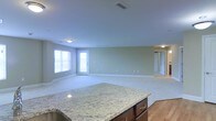This single-floor space is designed with an open-concept layout that seamlessly connects the kitchen and living areas, creating a bright and airy atmosphere. The kitchen features a generously sized island topped with elegant light-toned granite countertops with subtle veining, complemented by rich dark cabinetry beneath — a functional and visually striking centerpiece. The transition from the kitchen's warm hardwood flooring into the carpeted living area adds a comfortable and welcoming feel underfoot. The expansive living space is finished with soft neutral green-gray walls and white baseboards, lending the room a clean, understated aesthetic. Multiple windows along the far wall invite an abundance of natural light, enhancing the open and spacious feel of the room. Ceiling-mounted light fixtures provide additional illumination throughout. A doorway visible at the rear of the space leads to additional rooms, suggesting further utility and flow beyond the main living area. Overall, the space presents a practical yet polished environment well-suited for everyday living, with thoughtful material choices and a cohesive, neutral color palette throughout.