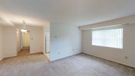 This single-floor space offers a clean, functional layout designed to maximize usability. The main living area is spacious and open, featuring neutral-toned wall-to-wall carpeting that adds warmth underfoot. Bright white walls provide a blank canvas, while a large window dressed with vertical blinds allows natural light to filter into the room, creating an airy atmosphere. Structural beam detailing along the ceiling adds a subtle architectural element to the otherwise minimalist design.

Adjacent to the living area, a transition to hard flooring marks the entry into the kitchen, which is visible through an open passageway. The kitchen is equipped with a full-size refrigerator, and the lighter flooring in this zone provides a practical, easy-to-clean surface. The layout facilitates a natural flow between the living and kitchen areas, making the space feel connected and cohesive.

Overall, the unit presents a straightforward, well-maintained interior with a neutral color palette throughout. The simple, understated design provides a versatile foundation that can accommodate a variety of furnishing and décor styles.