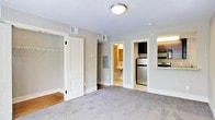 This single-floor unit offers a straightforward and functional layout designed to maximize usability within a modest footprint. The main living area is finished with wall-to-wall carpet in a neutral gray tone, providing a comfortable underfoot feel, while the walls are painted in a warm gray that lends a clean, understated aesthetic throughout. Overhead, flush-mount ceiling lighting keeps the space well-illuminated.

A built-in closet with white panel doors and interior coat hooks is conveniently located within the main living area, offering practical storage. Adjacent to it, an electrical panel is recessed into the wall. A hallway leads to a full bathroom, which features a pedestal sink and light-toned flooring, visible through the open doorway.

The kitchen is accessible from the main living space and is equipped with dark wood cabinetry, a stainless steel refrigerator, and a range with an overhead microwave. A countertop pass-through connects the kitchen to the living area, enhancing the sense of openness. Hardwood flooring transitions into the kitchen area, distinguishing it from the carpeted living space. Overall, the unit presents a clean, move-in-ready environment suited for efficient single-floor living.