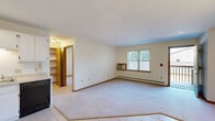 This single-floor residential unit offers a practical and comfortable layout designed for everyday living. The open living area features neutral-toned carpet, a wall-mounted air conditioning unit, and a baseboard heating system, with a large window fitted with vertical blinds that allows natural light to fill the space and offers views of the surrounding greenery. The kitchen is efficiently arranged with ample white cabinetry, light countertops, tile flooring, and a full suite of appliances including a refrigerator, electric range, built-in microwave, and dishwasher, complemented by a track lighting fixture overhead. A distinctive teal six-panel door leads from the living area to a private balcony, which overlooks a peaceful landscape of tall pine trees and manicured grounds. The bedrooms are carpeted and well-proportioned, each with vertical blind-covered windows and baseboard heating; one bedroom features a ceiling fan with light fixture. A generously sized walk-in closet provides ample storage with multiple wood shelving units and hanging rods. The unit's warm wood trim throughout ties the interior together, creating a cohesive and inviting atmosphere across every room.