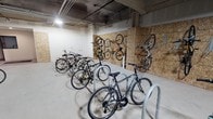Located conveniently near the elevator entrance, this dedicated bicycle storage room is designed to accommodate the needs of cycling residents. The space features a practical combination of wall-mounted vertical bike racks and freestanding floor-level hoops, maximizing storage capacity while keeping the area organized and accessible. The walls are lined with oriented strand board (OSB) panels, providing a sturdy surface for the wall-mounted hooks that allow bikes to be stored vertically, saving floor space. The exposed concrete ceiling reveals overhead piping and conduits, reflecting the room's utilitarian character. The polished concrete floor is easy to maintain and contributes to the clean, functional aesthetic of the space. Overhead lighting ensures visibility throughout. The room also appears to include an additional smaller area, visible in select images, with wire shelving and hanging storage — suggesting a supplementary utility or changing space nearby. Louvered ventilation panels provide airflow, keeping the environment well-ventilated. Overall, this space delivers a thoughtfully designed, secure, and efficient solution for bicycle storage, catering to an active, urban lifestyle.