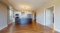 This single-floor residence offers a thoughtfully designed open-concept layout that seamlessly connects the kitchen and living areas. The kitchen is a standout feature, showcasing white shaker-style cabinetry with glass-front upper cabinets, granite countertops, a subway tile backsplash, and a full suite of stainless steel appliances including a range, over-the-range microwave, and refrigerator. A dark-finished island with a granite top provides additional prep space and a natural visual anchor to the room. Warm hardwood flooring flows throughout the main living areas, complemented by soft, neutral-toned walls and abundant natural light. A pendant chandelier adds a decorative focal point above the kitchen island. The living area opens to a private balcony through a sliding glass door, offering outdoor views of surrounding trees and greenery. The bedroom features plush carpet flooring, a large window with blinds, and a clean, spacious layout. The bathroom is finished with travertine-style tile flooring, a dark wood vanity with granite countertop, an undermount sink, and a full soaking tub with a tiled surround. The overall design is modern, cohesive, and move-in ready.