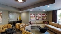 This single-floor space showcases a thoughtfully designed interior that blends contemporary style with warm, inviting elements. The lounge area is anchored by a large, neutral-toned sectional sofa adorned with richly colored accent pillows in deep plum and gold tones, complemented by a pair of tufted caramel leather armchairs that add a classic yet modern touch. A sleek, dark round coffee table with a woven decorative tray serves as a functional centerpiece, tying the seating arrangement together.

The walls are adorned with a striking triptych of abstract artwork featuring bold tones of burgundy, gold, and cream, adding visual depth and personality to the space. Recessed lighting is thoughtfully placed throughout the ceiling, providing even, ambient illumination, while a distinctive sunburst ceiling fixture adds a sculptural focal point overhead.

Along one wall, a dark console or credenza is flanked by tall, metallic gold decorative accents, enhancing the room's upscale aesthetic. A textured, reflective wall panel in the background introduces additional visual interest. The overall ambiance is polished and refined, making this space well-suited for social gatherings or relaxed lounging.